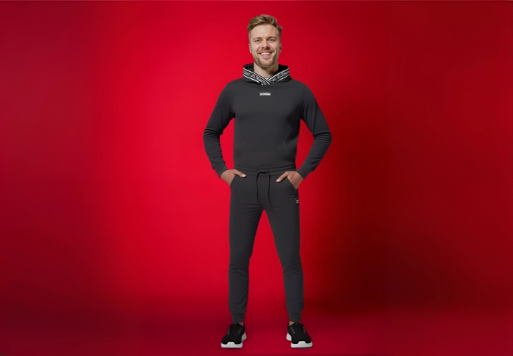 Een model staat in een strakke zwarte hoodie en bijpassende joggingbroek tegen een levendige rode achtergrond, met de handen in de zakken en sportieve sneakers aan.