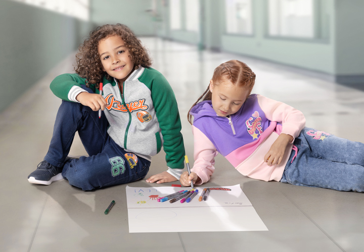 Twee kinderen zitten op een tegelvloer en tekenen met gekleurde stiften op een groot vel papier.