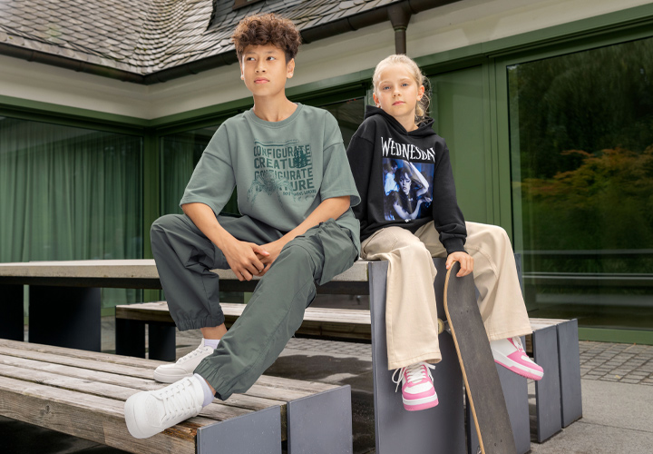 Een jongen in een oversized groen T-shirt en groene broek zit op een houten bank naast een meisje in een zwarte hoodie en beige broek, met een skateboard in haar hand.