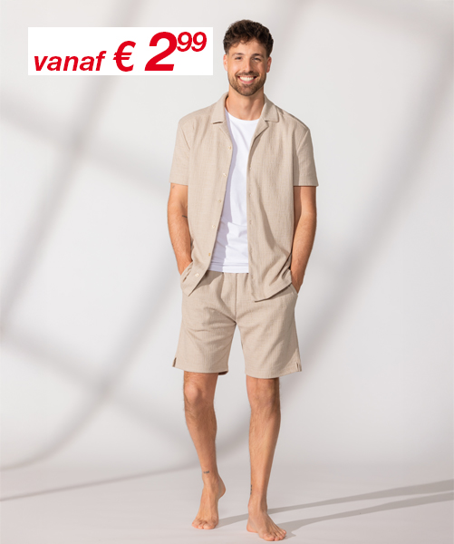 Een man staat blootsvoets, draagt een beige overhemd met korte mouwen en bijpassende shorts, met zijn handen nonchalant in de zakken – tegen een lichte achtergrond.