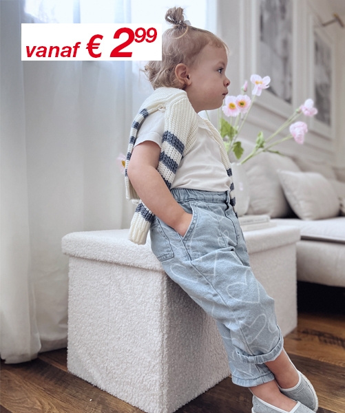 Een kind leunt tegen een witte poef, gekleed in een gestreepte trui en lichtblauwe broek, met op de achtergrond een moderne woonkamer en verse bloemen.