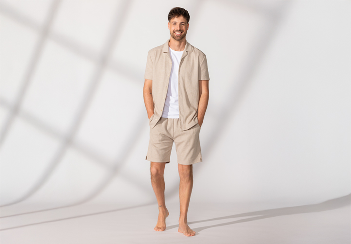 Een man staat blootsvoets, draagt een beige overhemd met korte mouwen en bijpassende shorts, met zijn handen nonchalant in de zakken – tegen een lichte achtergrond.