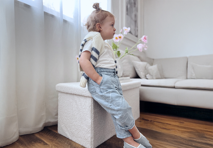 Een kind leunt tegen een witte poef, gekleed in een gestreepte trui en lichtblauwe broek, met op de achtergrond een moderne woonkamer en verse bloemen.