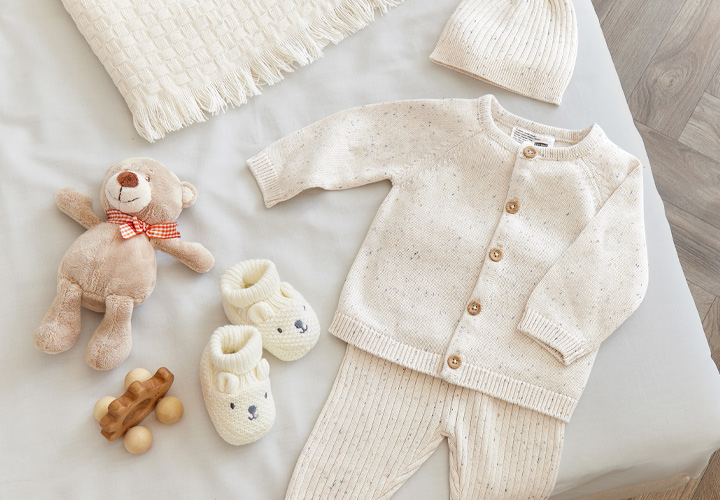 Gezellige babyuitzet met een gebreid vest, bijpassende broek, een muts, slofjes, een teddybeer en houten speelgoed op een zachte deken.